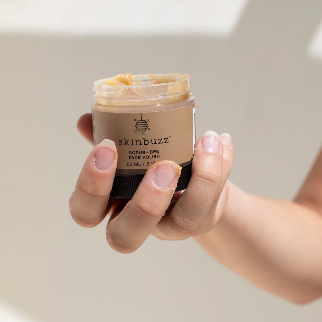 
                  
                    A hand holding an open jar of skinbuzz Acne Prone Face Polish, showing the beige cream product inside and the brown jar, set against a warm, softly lit beige background.
                  
                