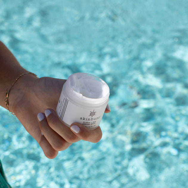 
                  
                    A person in a teal bikini holds an open white jar of skin cream over a bright turquoise swimming pool background.
                  
                