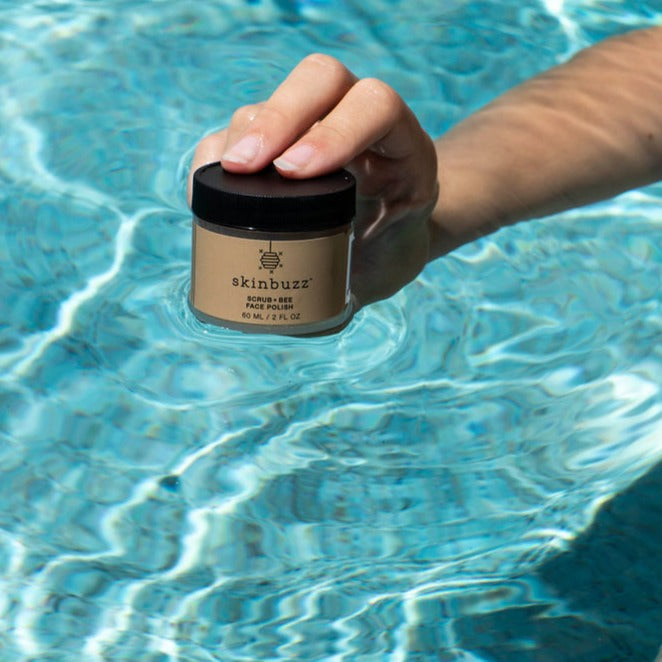 
                  
                    A hand holding a small jar of skinbuzz clay mask above rippling blue pool water.
                  
                